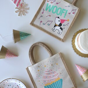 May include: Two small burlap gift bags with handles. One bag features a cartoon dog wearing a party hat with the word "WOOF!" in green letters. The other bag features a cartoon cupcake with pink frosting and sprinkles. Both bags have a brown burlap trim.