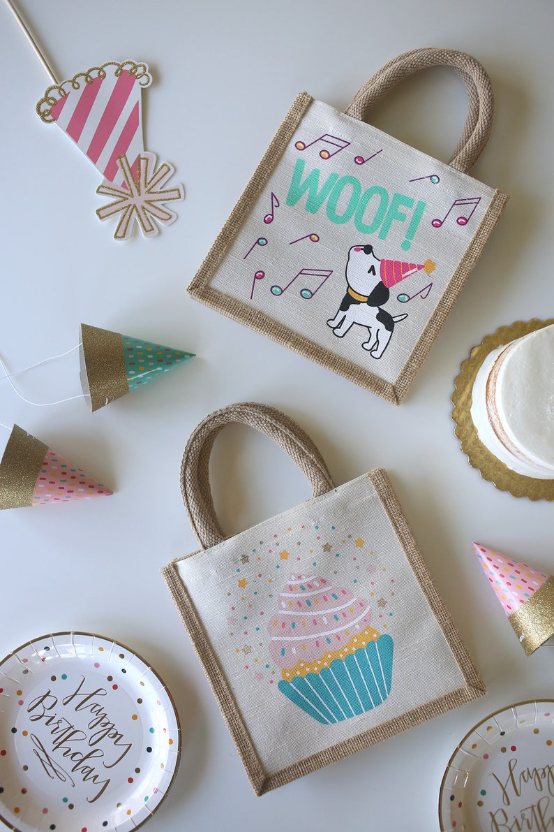 May include: Two small burlap gift bags with handles. One bag features a cartoon dog wearing a party hat with the word "WOOF!" in green letters. The other bag features a cartoon cupcake with pink frosting and sprinkles. Both bags have a brown burlap trim.