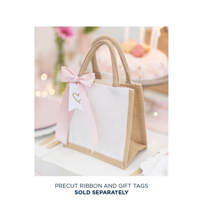 May include: A small white burlap tote bag with a pink ribbon and a gift tag with a gold heart. The bag has brown burlap trim.
