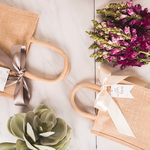 May include: Two small, tan burlap tote bags with rope handles and satin ribbon bows. One has a tag with heart shapes, the other says "with love." A bouquet of purple flowers and a succulent are also present.