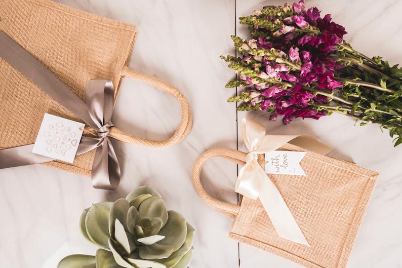 May include: Two small, tan burlap tote bags with rope handles and satin ribbon bows. One has a tag with heart shapes, the other says "with love." A bouquet of purple flowers and a succulent are also present.