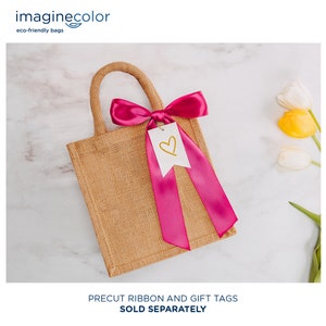 May include: A small, brown burlap tote bag with a pink satin ribbon tied in a bow and a white gift tag with a gold heart. The bag is on a white marble surface.