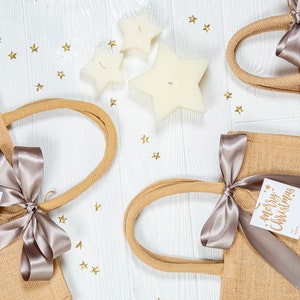 May include: Four burlap tote bags with tan handles and silver ribbon bows, each adorned with a white tag that reads "Merry Christmas" in gold lettering. Three star-shaped candles and gold star confetti are scattered on a white wooden surface.