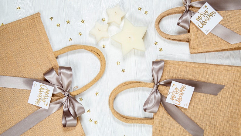 May include: Four burlap tote bags with tan handles and silver ribbon bows, each adorned with a white tag that reads "Merry Christmas" in gold lettering. Three star-shaped candles and gold star confetti are scattered on a white wooden surface.