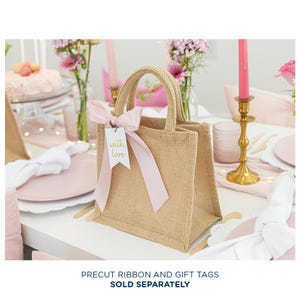May include: A small, brown burlap tote bag with a pink ribbon and a white gift tag that says "with love" in gold lettering. The bag is sitting on a white table with pink and white decorations.