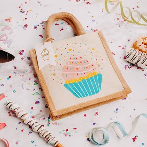 May include: A small, beige hessian tote bag with a colourful cupcake design. The bag has a handle and a tag that says "Happy Birthday". The bag is surrounded by confetti and colourful streamers.