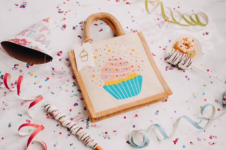 May include: A small, beige hessian tote bag with a colourful cupcake design. The bag has a handle and a tag that says "Happy Birthday". The bag is surrounded by confetti and colourful streamers.