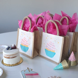 May include: A set of six small burlap gift bags with pink tissue paper, each featuring a cupcake design with sprinkles and stars. The bags are on a white table with a cake, party hats, and other decorations.