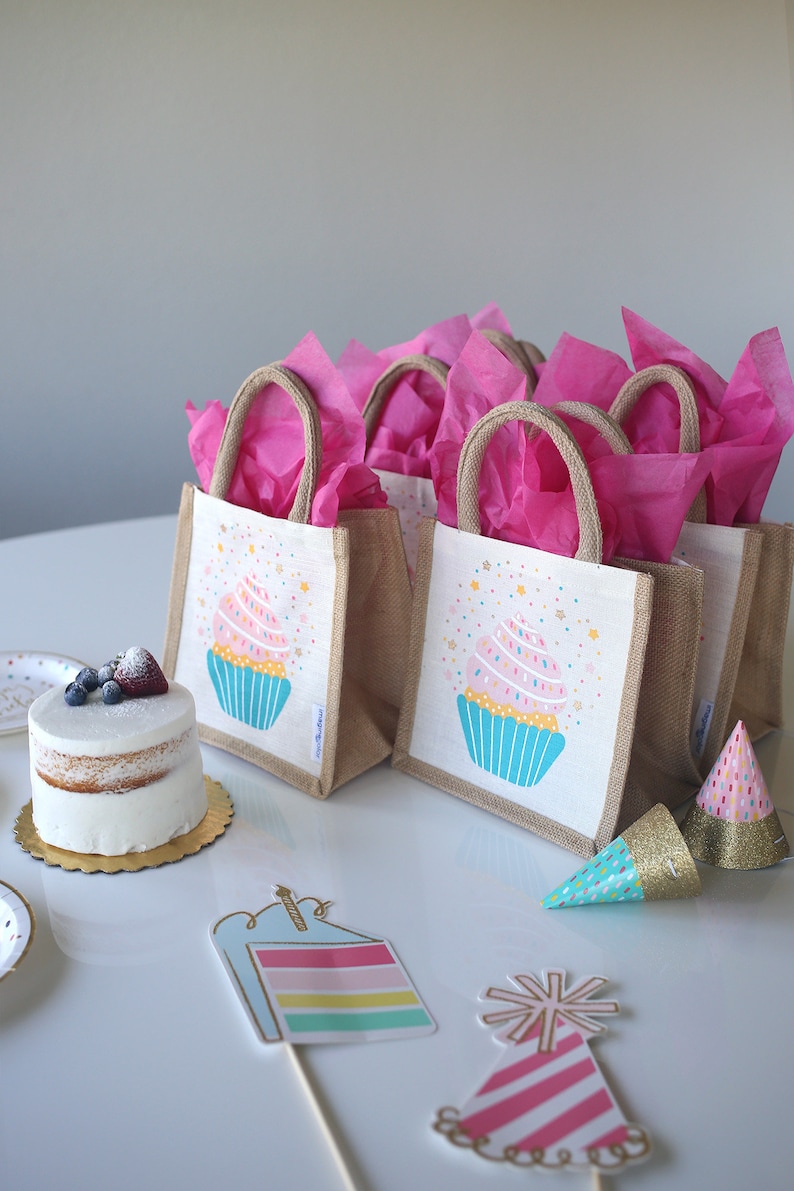 May include: A set of six small burlap gift bags with pink tissue paper, each featuring a cupcake design with sprinkles and stars. The bags are on a white table with a cake, party hats, and other decorations.