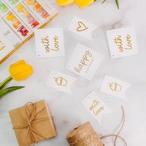May include: A gift box wrapped in brown paper with twine, a spool of twine, and several white gift tags with gold lettering. The tags say "happy birthday", "with love", and a heart. There are also two tags with a cupcake illustration.