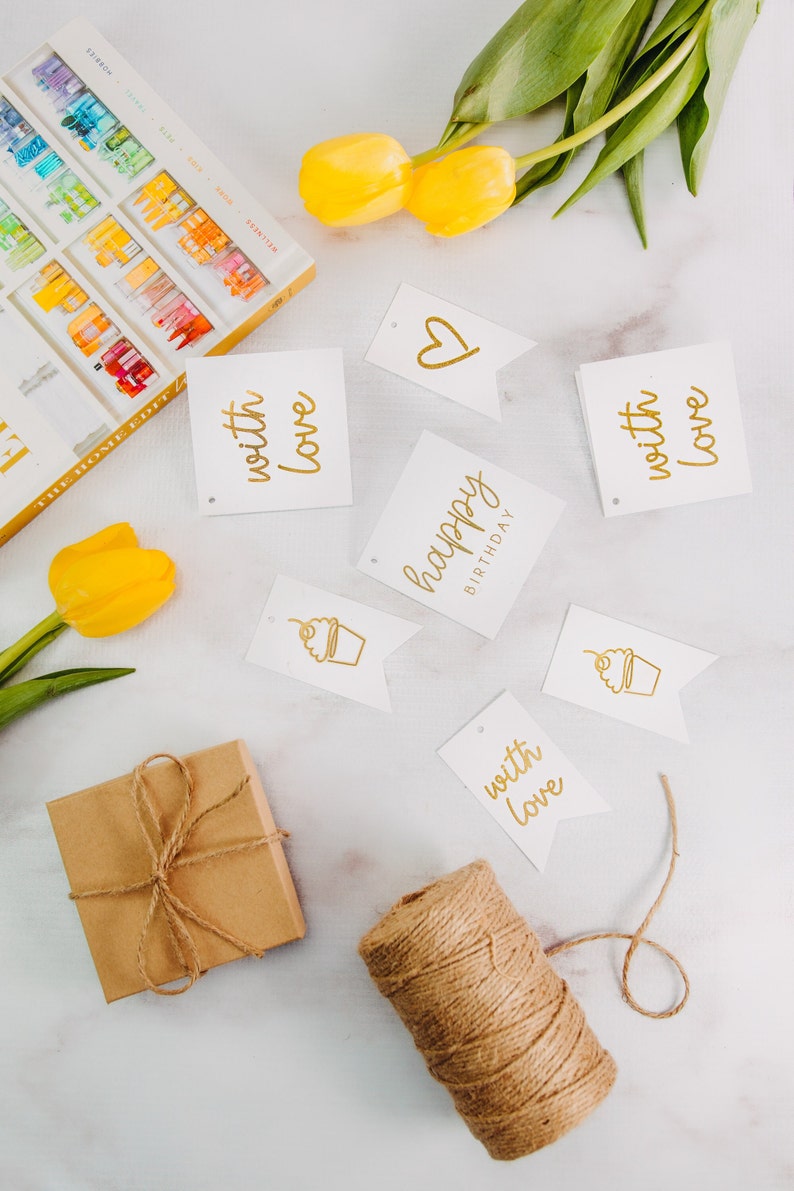 May include: A gift box wrapped in brown paper with twine, a spool of twine, and several white gift tags with gold lettering. The tags say "happy birthday", "with love", and a heart. There are also two tags with a cupcake illustration.