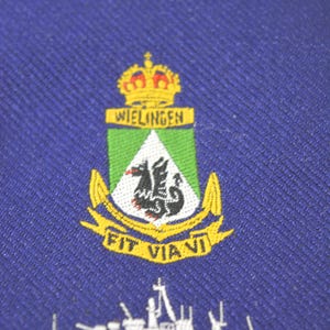 Könnte beinhalten: Marineblaue Aufnäher mit Wappen, Krone, dem Wort "WIELINGEN" und einem schwarzen Drachen. Darunter der Schriftzug "FIT VIANI" und eine weiße Silhouette eines Schiffs.