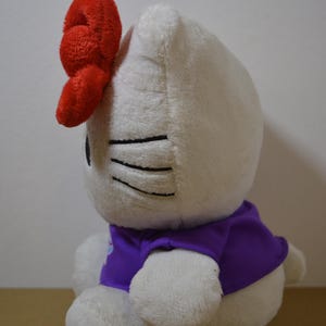 May include: A white plush toy cat with a large red bow on its head. The cat wears a purple shirt. Black lines represent the cat's eyes and whiskers. The toy is soft and cuddly.