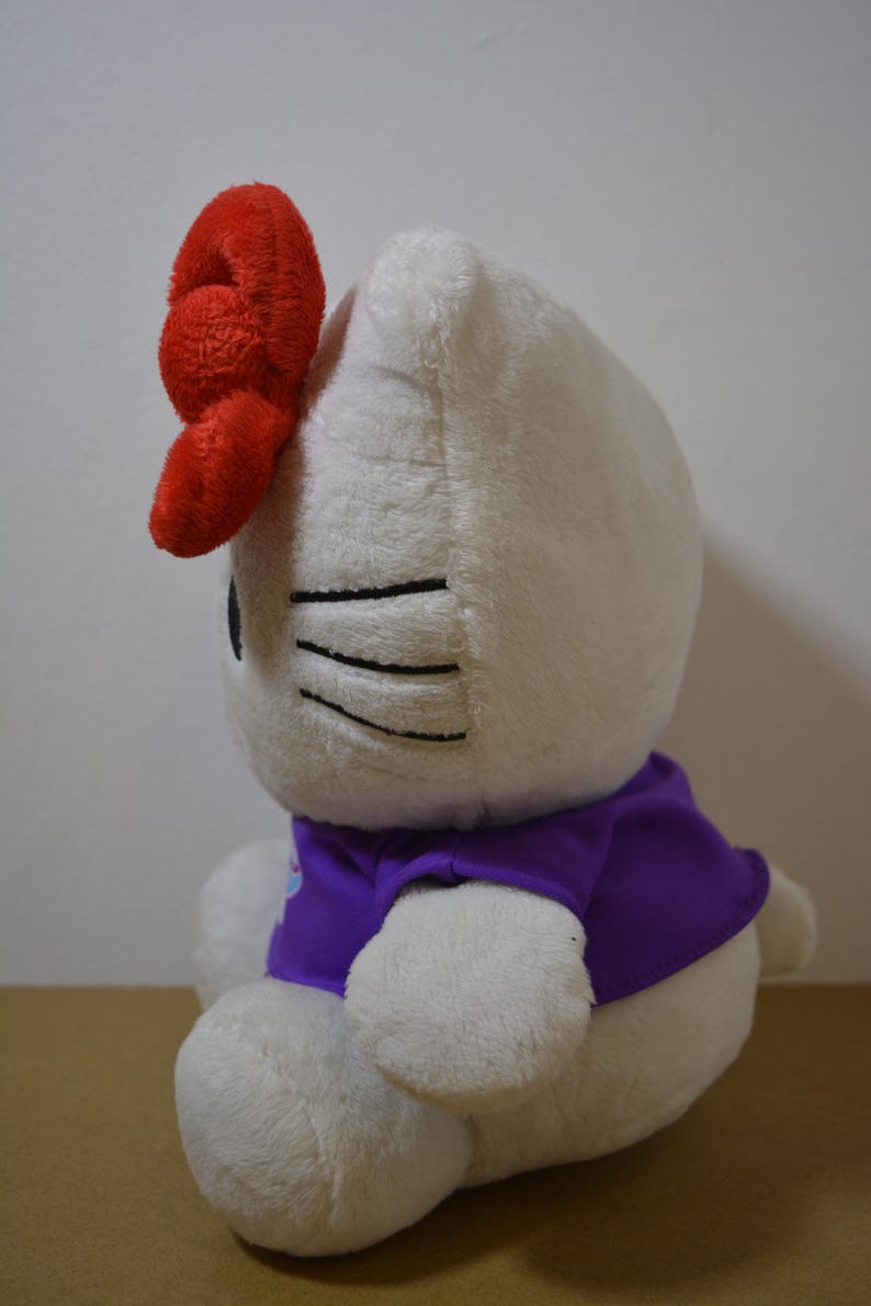 May include: A white plush toy cat with a large red bow on its head. The cat wears a purple shirt. Black lines represent the cat's eyes and whiskers. The toy is soft and cuddly.