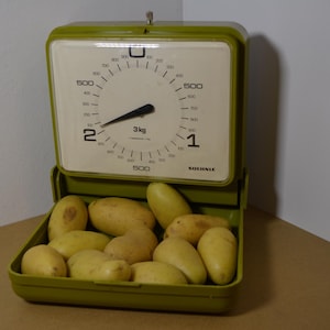 May include: A vintage olive green Soehnle kitchen scale with a square face and a circular analog dial measuring up to 3kg. The scale features a hinged base tray filled with a collection of raw, light brown potatoes. The dial displays black markings.