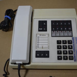 Może przedstawiać: Kremowy, zabytkowy telefon z białą słuchawką i czarnymi przyciskami. Telefon posiada klawiaturę numeryczną, przyciski funkcyjne i małą szczelinę na karty. Naklejka z tekstem znajduje się po prawej stronie, a przewód jest zabezpieczony niebieską opaską.