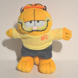 May include: A plush orange Garfield toy wearing a yellow shirt with a paw print and blue jeans. The toy has a black headband and a moustache.
