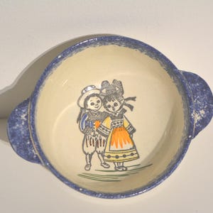 Könnte beinhalten: Eine weiße Keramikschüssel mit blau-weißem Rand. Die Schüssel zeigt ein handgemaltes Design von zwei Kindern in traditioneller Kleidung. Die Schüssel hat zwei Griffe.