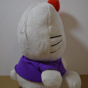 May include: A white plush toy with a red pom-pom and a purple shirt. The toy has black lines for eyes and a white string. The toy is sitting on a brown surface.
