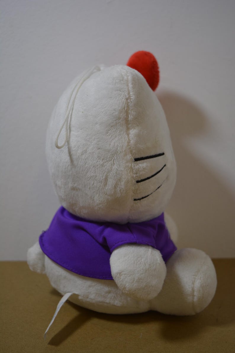May include: A white plush toy with a red pom-pom and a purple shirt. The toy has black lines for eyes and a white string. The toy is sitting on a brown surface.