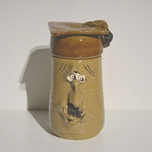May include: A ceramic mug with a face and a graduation cap lid. The mug is light brown with a darker brown glaze. The face has a large nose, small eyes, and a moustache.