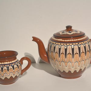 Peut inclure: Une théière en céramique marron avec un pot à crème assorti. Les deux pièces présentent un motif décoratif de dessins verticaux noirs, blancs et orange. La théière a un couvercle et un bec courbé. Le pot à crème a une anse et un bec verseur.
