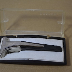 Può includere: Un rasoio di sicurezza vintage con testina in metallo argentato e manico nero testurizzato, riposto in una custodia rettangolare in plastica nera con coperchio trasparente incernierato. La parola Shick è impressa sul coperchio in plastica.