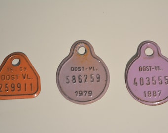 May include: Three vintage metal tags in orange, pink, and lavender. The tags have a hole at the top and feature stamped numbers and text, including "OOST VL." and years like 1969, 1979, and 1987. The orange tag is triangular.