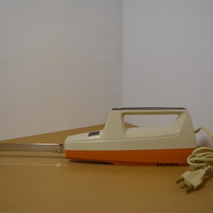 May include: An electric carving knife with a cream and orange body and a serrated stainless steel blade. The knife has a handle and a power cord with a plug. The knife is designed for slicing meats and other foods.
