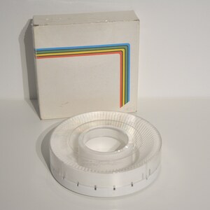 Può includere: Un carosello per proiettore di diapositive in plastica bianca con un coperchio in plastica trasparente. Il carosello è vuoto e ha una serie di fessure attorno al bordo. Il carosello è appoggiato su una superficie bianca. La scatola del carosello è bianca con un design a strisce arcobaleno.