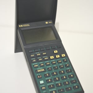 Peut inclure: Une calculatrice graphique Hewlett Packard 38G noire et verte avec un couvercle de protection. La calculatrice possède un écran, de nombreux boutons et le logo Hewlett Packard. Le couvercle est ouvert, révélant l'écran et le clavier.