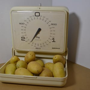 Op de afbeelding: Een crèmekleurige Soehnle keukenweegschaal met een vierkant analoog display met een capaciteit van 3 kg. De weegschaal heeft een scharnierend, geïntegreerd plateau aan de onderkant gevuld met diverse rauwe, lichtbruine aardappelen.