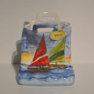 May include: A ceramic vase with a handle, painted with a nautical scene. The design features two sailboats with red and green sails on a blue sea, under a blue sky. A yellow sticker is affixed to the vase.