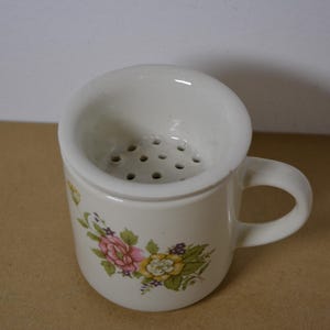 Può includere: Una tazza in ceramica bianca dotata di un colino integrato con molteplici fori di drenaggio. L'esterno della tazza è decorato con un motivo floreale composto da fiori rosa, gialli e verdi con piccoli accenti viola.