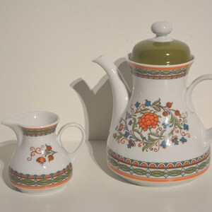 May include: A white ceramic teapot and creamer set with a floral pattern in shades of orange, green, and blue. The teapot has a green lid and a floral design on the body. The creamer has a matching floral design.
