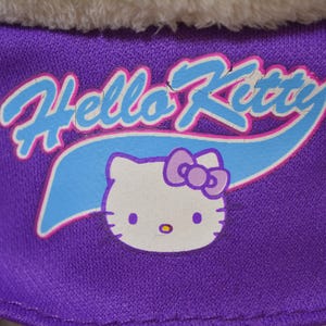 May include: Close-up of a purple fabric with the words "Hello Kitty" in white and light blue, with a cartoon image of Hello Kitty. The image features a white cat face with a purple bow and yellow nose.