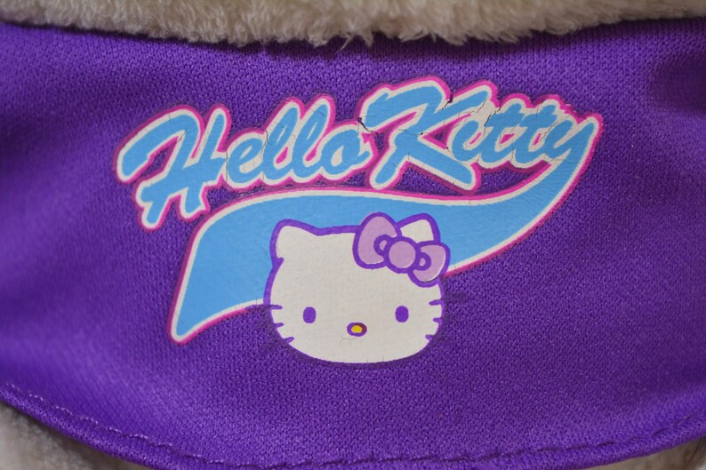 May include: Close-up of a purple fabric with the words "Hello Kitty" in white and light blue, with a cartoon image of Hello Kitty. The image features a white cat face with a purple bow and yellow nose.