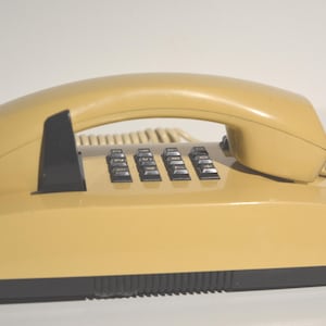 Könnte beinhalten: Ein beigefarbenes Wählscheibentelefon mit einem schwarzen Tastenfeld und einem beigefarbenen Hörer. Das Telefon hat ein Spiralkabel.
