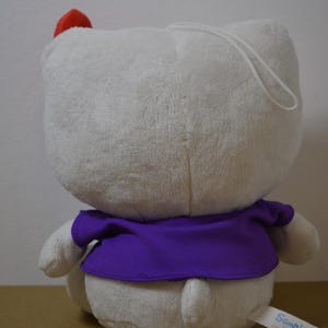 May include: A plush white character with a red bow and purple shirt. The stuffed animal has a loop for hanging and a tag that says "Sanrio". The toy is soft and fluffy.