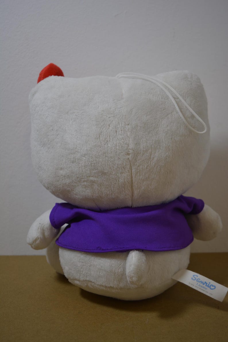 May include: A plush white character with a red bow and purple shirt. The stuffed animal has a loop for hanging and a tag that says "Sanrio". The toy is soft and fluffy.
