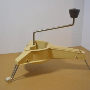 May include: A vintage, cream-colored kitchen tool with a crank handle and three legs. The handle is dark brown, and the legs are silver with white tips. The tool appears to be made of metal and plastic.