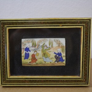 May include: A framed miniature painting depicting figures in colorful robes. The painting is set against a black background within a decorative frame with gold and brown accents. The artwork showcases a scene with multiple figures in a landscape.