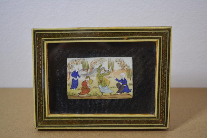 May include: A framed miniature painting depicting figures in colorful robes. The painting is set against a black background within a decorative frame with gold and brown accents. The artwork showcases a scene with multiple figures in a landscape.