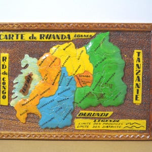 May include: A framed, colorful map of Rwanda, featuring the country's borders and internal regions. The map is labeled with the text "CARTE du RWANDA" and includes the names of neighboring countries and regions. The frame is a dark brown color.
