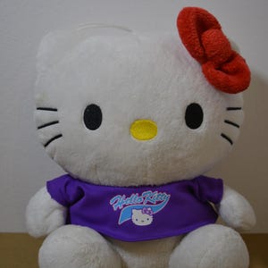 May include: A white Hello Kitty plush toy with a red bow, black eyes, and a yellow nose. The plush is wearing a purple shirt with the Hello Kitty logo. The toy is sitting on a brown surface.