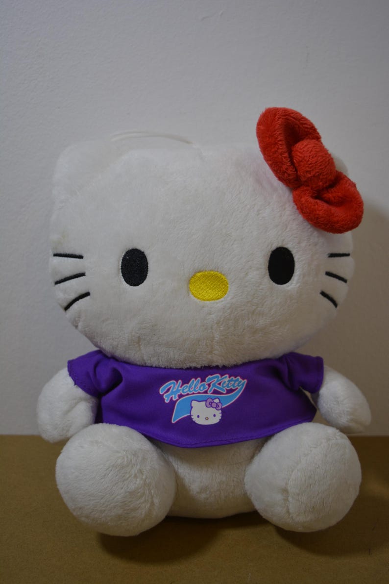 May include: A white Hello Kitty plush toy with a red bow, black eyes, and a yellow nose. The plush is wearing a purple shirt with the Hello Kitty logo. The toy is sitting on a brown surface.