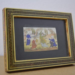 May include: A framed miniature painting depicting figures in colorful robes in a landscape scene. The frame is rectangular with a decorative border in shades of gold and brown. The artwork is set against a black background.