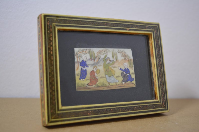 May include: A framed miniature painting depicting figures in colorful robes in a landscape scene. The frame is rectangular with a decorative border in shades of gold and brown. The artwork is set against a black background.