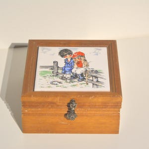 May include: A wooden box with a hinged lid and a metal latch. The box has a ceramic tile on the front with a cartoon image of two children sitting on a fence. The children are wearing blue and red clothing.