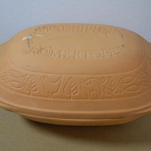 Pode incluir: Uma assadeira oval de barro terracota com tampa. A tampa apresenta o texto em relevo ROMERTOPF e uma borda decorativa com motivos alternados de peixes, folhas e porcos moldados na superfície da argila natural.
